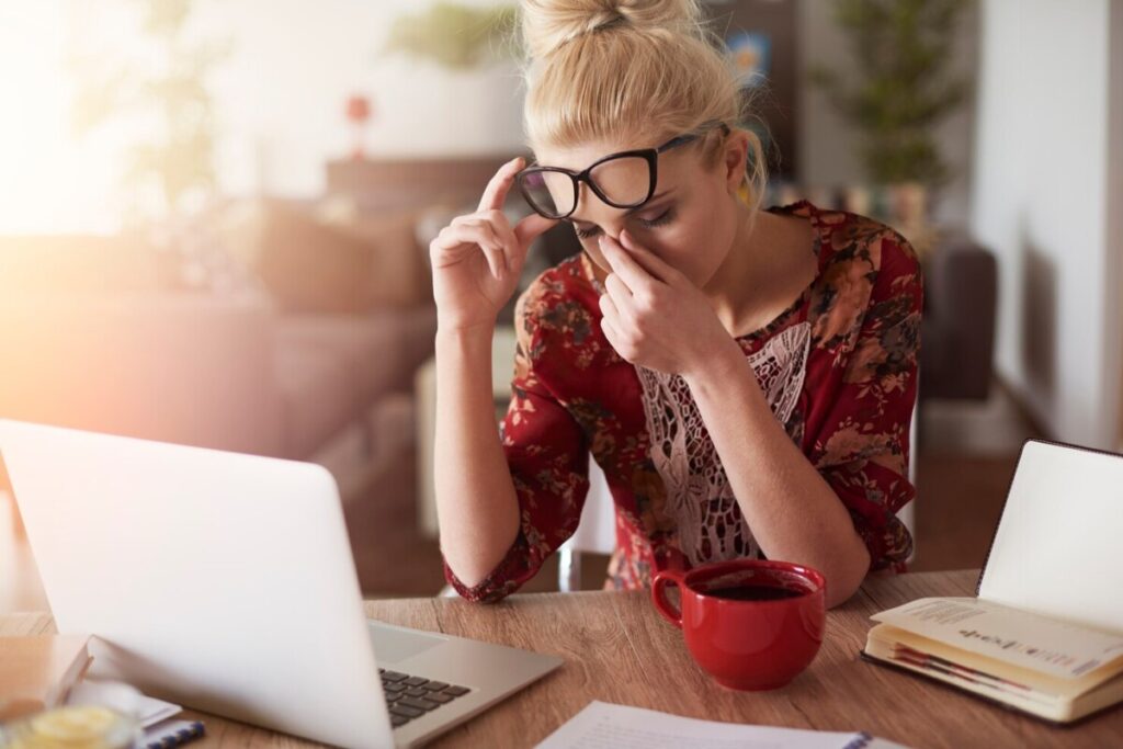 En accord avec l'accent mis par le site sur l'Ashwagandha, l'image met en valeur cette plante remarquable comme remède potentiel contre le stress. Assise à un bureau avec un ordinateur portable ouvert, la femme aux cheveux blonds et à la tenue fleurie est photographiée tenant ses lunettes et se pinçant le nez dans un moment de contemplation du stress. Alors qu'une tasse rouge et des livres sont présents sur la table, c'est l'Ashwagandha qui se distingue comme une solution idéale pour favoriser le calme et l'équilibre.